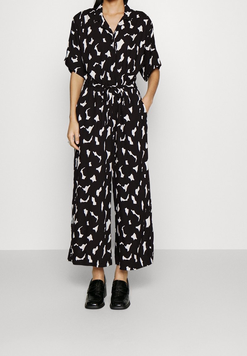 Combinaison noire avec des motifs abstraits blancs, manches courtes, un col et une taille nouée. Présente des jambes larges et des poches latérales. Associée à des chaussures noires.