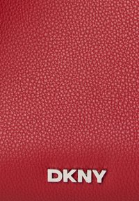 Rød struktureret læderoverflade med sølvmetallic DKNY mærkelogo centreret nær bunden.