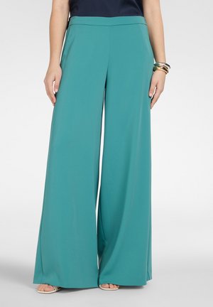 Kobieta ubrana w szerokie spodnie w kolorze morskim, czarną bluzkę, złote i srebrne bransoletki oraz białe sandały z odkrytymi palcami, stojąca na tle jednolitego tła.