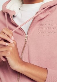 Manos ajustando el cordón de una sudadera con cremallera rosa, con forro de lana blanca y el texto bordado "Superdry Premium" en el pecho.