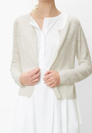 Kvinde iført en lysebeige tekstureret cardigan over en hvid knapklædt kjole, der lukker cardiganens forreste knapper.