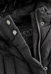 Veste d'hiver noire avec fermeture éclair argentée et boutons-pression, partiellement ouverte pour montrer l'étiquette en tissu et la bordure en fausse fourrure sur la capuche.