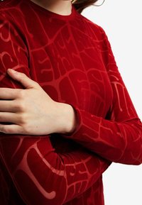 Personne croisant les bras, portant une chemise rouge à manches longues avec un motif texturé abstrait sur un fond blanc.