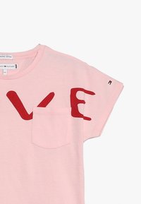 Camiseta rosa de manga corta hecha de algodón orgánico, con un gran gráfico rojo de "LOVE" y un pequeño bolsillo en el pecho. Branding de Tommy Hilfiger visible.