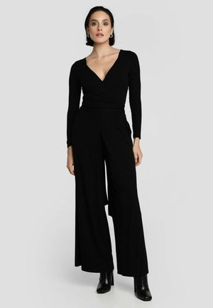 Donna che indossa una tuta nera a maniche lunghe con scollo a V, gambe larghe e stivali neri alla caviglia, in piedi contro uno sfondo chiaro e semplice.