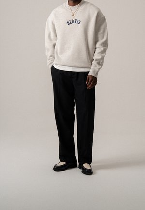 Mann trägt einen hellbeigen Pullover mit dem Text "BLKVIS", schwarze Hose, Loafers und goldene Halsketten vor einem schlichten Hintergrund.