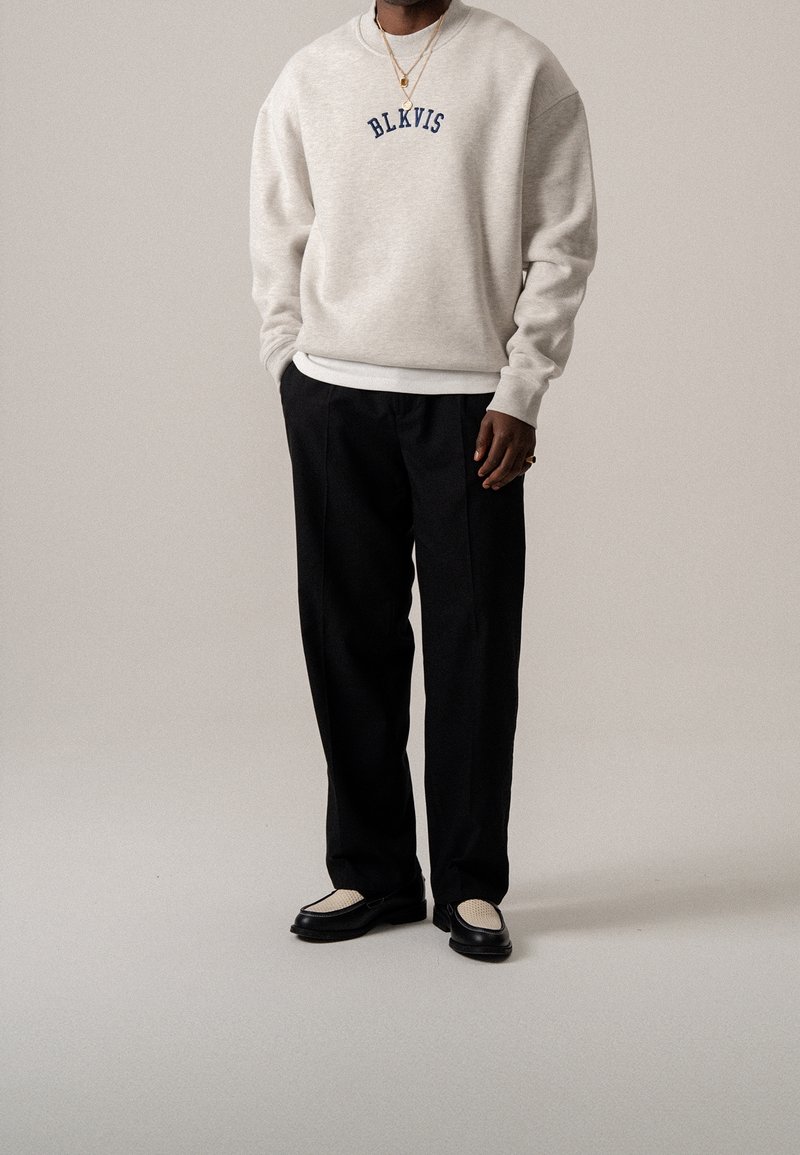 Homme portant un sweat-shirt beige clair avec le texte "BLKVIS", pantalon noir, mocassins et colliers dorés sur un fond uni.