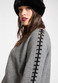 Grauer Strickpullover mit schwarzen Akzenten entlang der Ärmel. Verfügt über eine lockere Passform und strukturierte Strickdetails, kombiniert mit einem schwarzen Pelzhut.