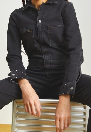 Person i sort denimjakke og bukser, siddende med benene fra hinanden på en metallisk ribbet overflade, hænder afslappede og hvilende fremad.