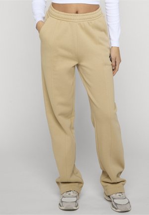 Persoon draagt beige sweatpants met rechte pijpen, handen in de zakken en witte sneakers, tegen een effen lichte achtergrond.