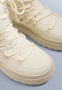 Kremowe wysokie botki z teksturowanymi gumowymi podeszwami, z krzyżującymi się sznurówkami i gładką syntetyczną cholewą z przeszyciami.
