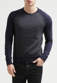 Hombre con un suéter azul marino de mangas largas y un patrón de cuadros en la parte frontal, combinado con jeans negros, con la mano en el bolsillo.
