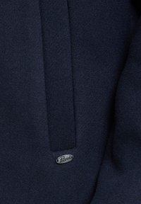 Marineblaues Wolljackett mit umgeschlagenem Kragen, strukturiertem Stoff und silbernem Logoabzeichen. Das Design betont klare Linien und eine klassische Silhouette.