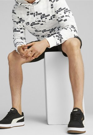 Man zit op een witte blok, draagt een witte Puma-hoodie met zwarte logo's, zwarte korte broek en zwarte Puma-sneakers met witte zolen.