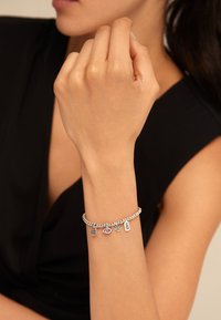 Pulsera de plata con detalles de cuentas, con charms variados que incluyen un ojo, una mano y etiquetas grabadas, llevada en una mano.