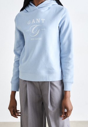 Osoba w jasnoniebieskiej bluzie z kapturem GANT z logo oraz luźnych, szarych spodni w prążki na białym tle.