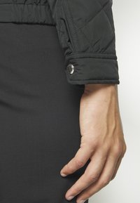 Hand with visible veins resting beside black quilted jacket sleeve with button detail, against black fabric background.