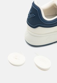 Sneaker med vit bas, marinblå texturerad hälkappa och två vita snören bredvid. Den har en upphöjd logotyp på hälen.