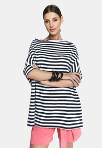 Pasiasty, oversize top w granatowo-białym materiale, z dekoltem karczkowym i krótkimi rękawami, zestawiony z różowymi szortami i czarną bransoletką.