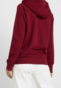 Person som bär en vinröd hoodie och vita byxor, sedd bakifrån mot en enkel ljus bakgrund.