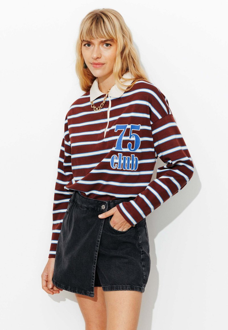 Polo à manches longues rayé marron et bleu avec un col et un logo sur le devant. Associé à une jupe en denim noire avec une fente sur le côté.
