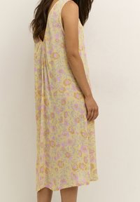 Kobieta w bezrękawnej żółtej sukience w różowy i pomarańczowy wzór kwiatowy, stojąca bokiem na tle jednolitego jasnego koloru.