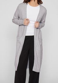 Jasnoszary kardigan o długim fasonie z otwartym przodem, z prążkowanymi mankietami i dwoma przednimi kieszeniami, noszony na białej bluzce i czarnych spodniach.