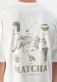 Bílý bavlněný tričko s zelenými ilustracemi matcha produktů a pumy, s nápisem "MATCHA The Puma Way" níže.