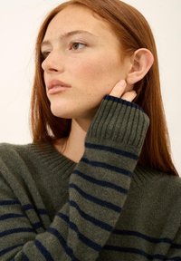 Zielony sweter w granatowe poziome paski, z prążkowanymi mankietami i szerokim dekoltem. Tekstura wydaje się miękka i dziana. Modelka dotyka swojego ucha.