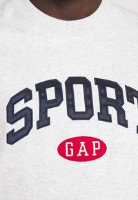 Sweat-shirt gris avec un grand texte "SPORT" bleu marine et un logo ovale rouge "GAP", fabriqué en coton doux avec une finition texturée.