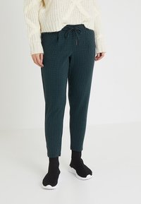 Pantalones de cuadros verdes con cintura ajustable, corte slim y textura sutil, combinados con zapatos negros sin cordones.