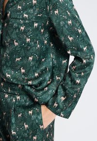 Camisa de pijama de satén verde con patrones de ciervos y árboles. Cuenta con un bolsillo de parche y un corte holgado. Textura suave con un brillo sutil.