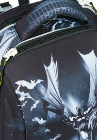 Schwarzer Rucksack mit grafischem Design eines Charakters mit Fledermäusen, mit Reißverschlüssen und einem strukturierten Griff. Akzente in Grau und Weiß.