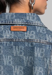 Jeansjacke mit blauer Waschung, geprägtem Logo-Muster und einem Lederanhänger mit "WRSTBHVR" auf der Rückseite. Akzente in Silber.