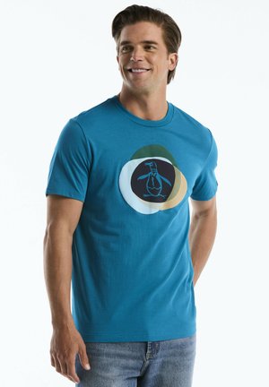 Man smiling wearing a blue short-sleeve t-shirt with a multicolored abstract penguin graphic on the chest and blue jeans.