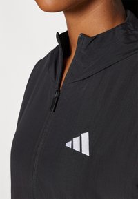 Svart zip-up jakke laget av lett stoff med høy krage. Har en hvit trekantet logo på brystet. Glatt tekstur.