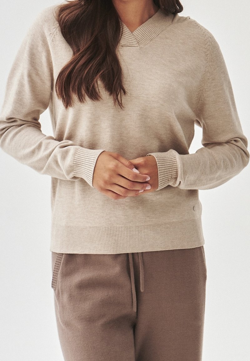 Femme portant un pull beige à col en V et un pantalon marron avec cordon de serrage, les mains légèrement jointes à la taille.