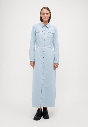 Lang, lyseblå denimkjole med lange ærmer, krage, brystlommer og knappestolpe foran. Bært med svarte sko.