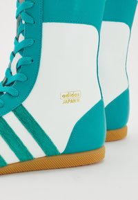 Tyrkysové a biele vysoké tenisky s textúrovanou zamatovou a hladkou kožou. Obsahujú zlaté logo "adidas JAPAN H" a vzorovanú gumenú podrážku.