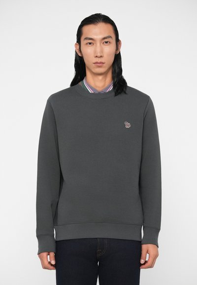 Un homme adulte aux longs cheveux noirs portant un sweat-shirt gris foncé par-dessus une chemise à rayures avec col et un jean noir, se tenant devant un fond uni.
