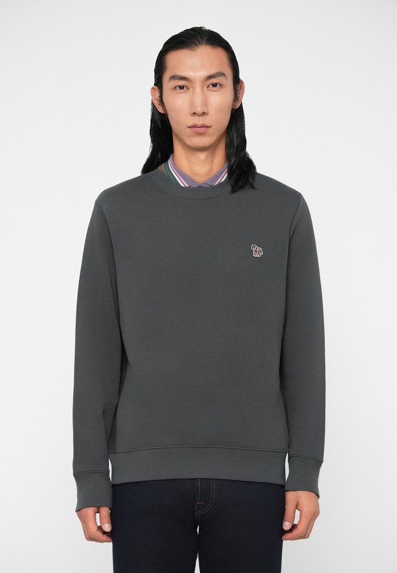 Suaugęs vyras su ilga juoda plauka, vilkintis tamsiai pilką megztinį per juostuotą marškinėlį ir tamsias džinsus, stovi prieš paprastą foną.