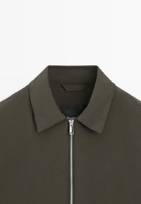 Mørkeolivengrøn jakke med spidst krave og central sølvlynlås på hvid baggrund.