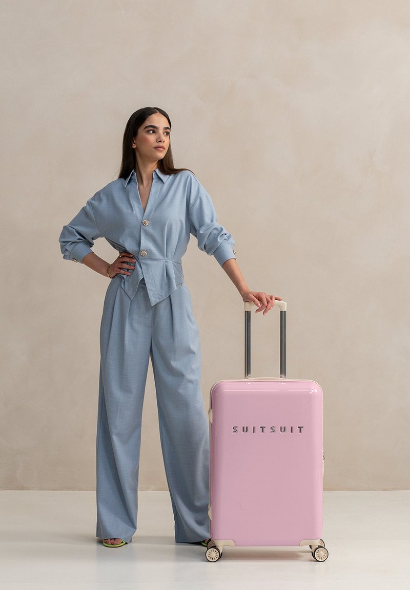 Lichtblauwe, knoopsluiting blouse en wijde broek met een glanzende roze koffer met zilveren 'SUITSUIT' logo en dubbele wielen.