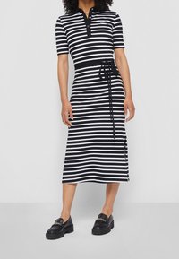 Nainen, jolla on yllä mustavalkoraidallinen midi-mekko lyhyillä hihoilla, nappikauluksella, vyötärönauha ja mustat loafersit kultaketjuilla.