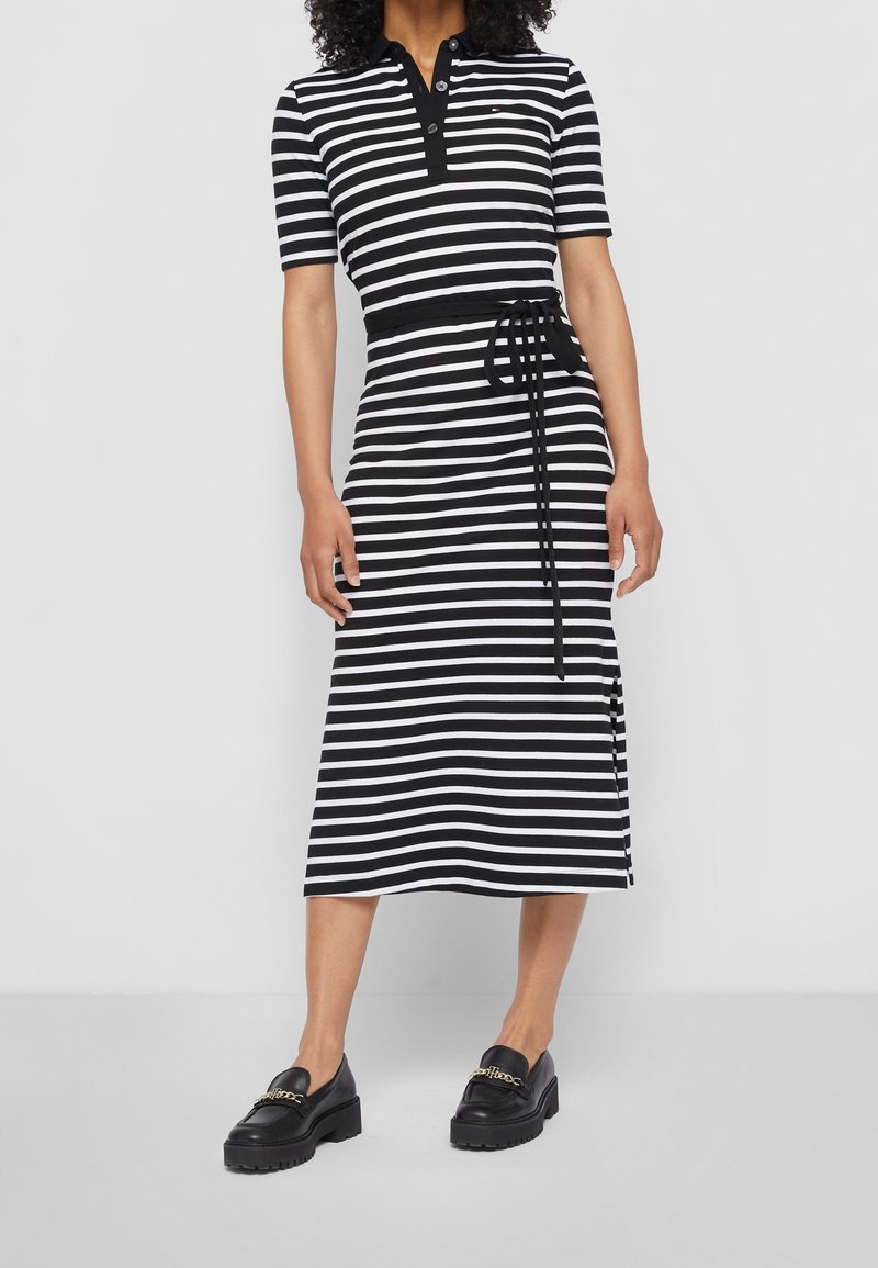 Nainen, jolla on yllä mustavalkoraidallinen midi-mekko lyhyillä hihoilla, nappikauluksella, vyötärönauha ja mustat loafersit kultaketjuilla.