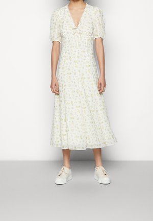 Mujer con un vestido midi blanco con un patrón floral amarillo, mangas abullonadas y zapatillas blancas de plataforma, de pie frente a un fondo liso.