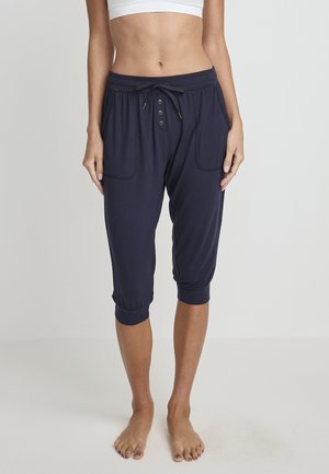 Frau steht barfuß und trägt marineblaue, verkürzte Hosen mit Kordelzug an der Taille und Knopfdetails, kombiniert mit einem weißen Crop-Top.