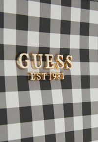 Väska med svartvit rutig mönster, prydd med en gyllene "GUESS"-logotyp och texten "EST 1981", med en slät textur.