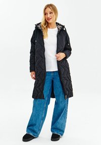 Prošívaný černý dlouhý kabát s vzorovaným interiérem, s kapucí, předním zipem a bočními kapsami, v kombinaci s volnými modrými džínami.