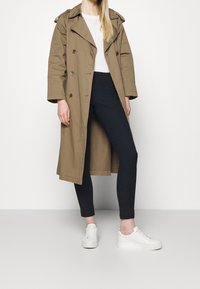 Tan trenchcoat med dubbelknäppning, över en vit topp, tillsammans med svarta leggings och vita sneakers mot en plain bakgrund.